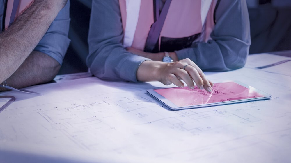 Die Person in der Sicherheitsweste zeigt auf ein Tablet, das auf Architekturplänen liegt.