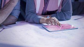 Die Person in der Sicherheitsweste zeigt auf ein Tablet, das auf Architekturplänen liegt.