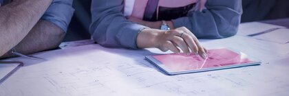 Die Person in der Sicherheitsweste zeigt auf ein Tablet, das auf Architekturplänen liegt.