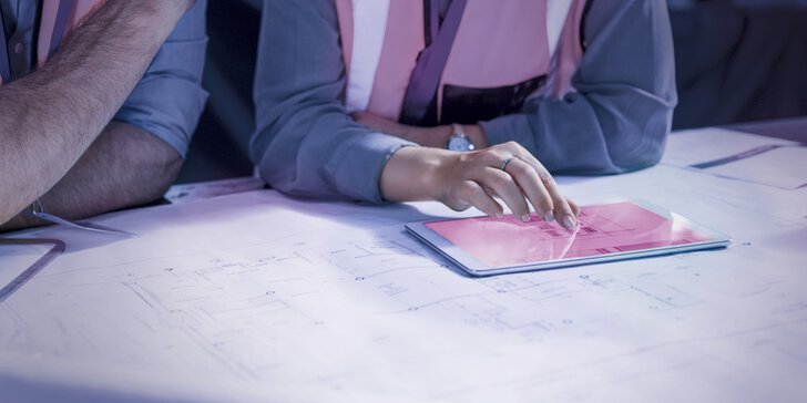 Die Person in der Sicherheitsweste zeigt auf ein Tablet, das auf Architekturplänen liegt.