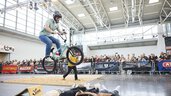 Ein Fahrrad Fahrer springt mit seinem BMX Rad über eine Absprungrampe über vier am Boden liegende Personen auf der Fahrrad-Parcours-Fläche auf der free-Messe