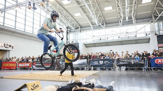 Ein Fahrrad Fahrer springt mit seinem BMX Rad über eine Absprungrampe über vier am Boden liegende Personen auf der Fahrrad-Parcours-Fläche auf der free-Messe
