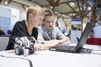 Zwei Jungen arbeiten in einem hellen, modernen Klassenzimmer an einem Laptop, auf dem ein kleiner Roboter steht.