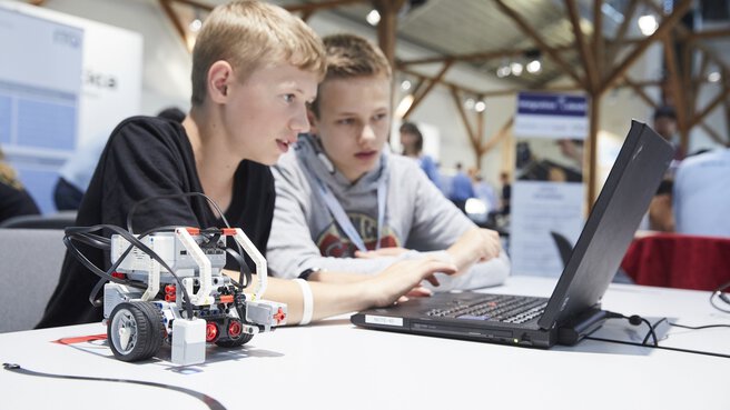 Two boys are working on a laptop with a small robot on it in a bright, modern classroom.