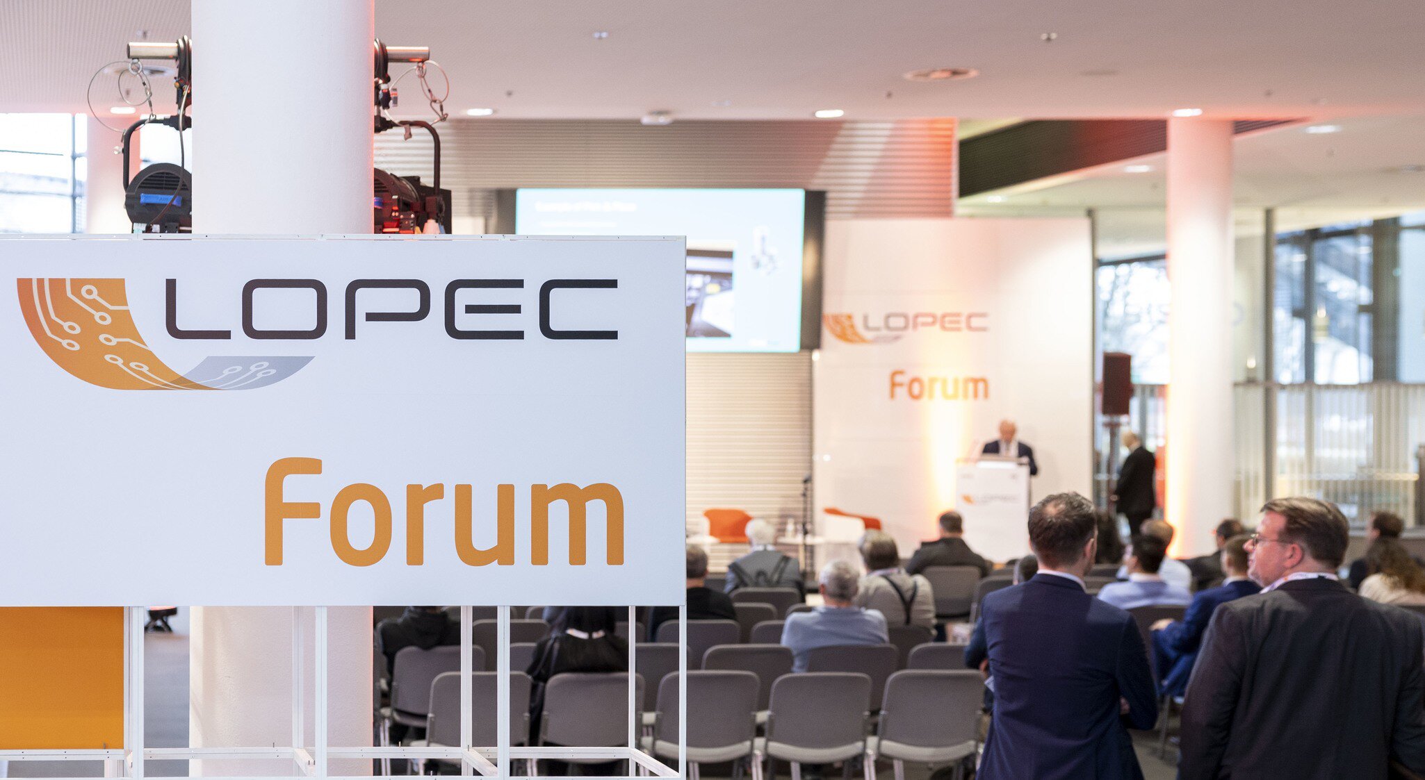Ein weißes Schild zum LOPEC Forum. Daneben unterhalten sich zwei Männer, während vor ihnen eine Präsentation gehalten wird.