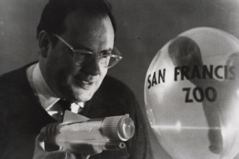 Black and white photo of a man aiming a laser at a balloon labeled 