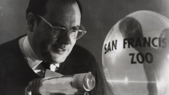 Black and white photo of a man aiming a laser at a balloon labeled 