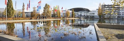 Blick über den Messesee der Messe München zum Westeingang, mit bunten Fahnen und herbstlichen Bäumen, die den Weg zum Eingang säumen.