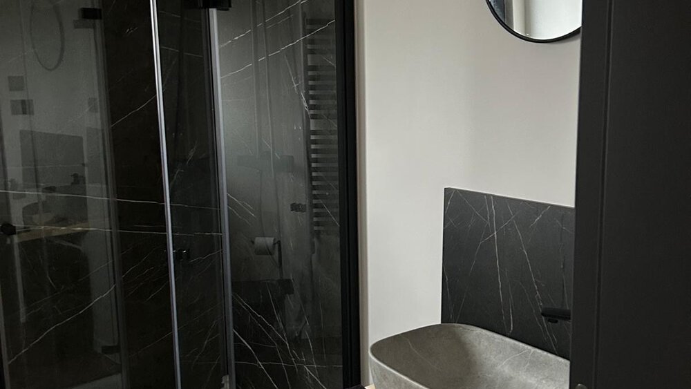 Modernes Badezimmer mit Glasdusche, rundem Spiegel, grauem Steinwaschbecken und schwarzen Akzenten.