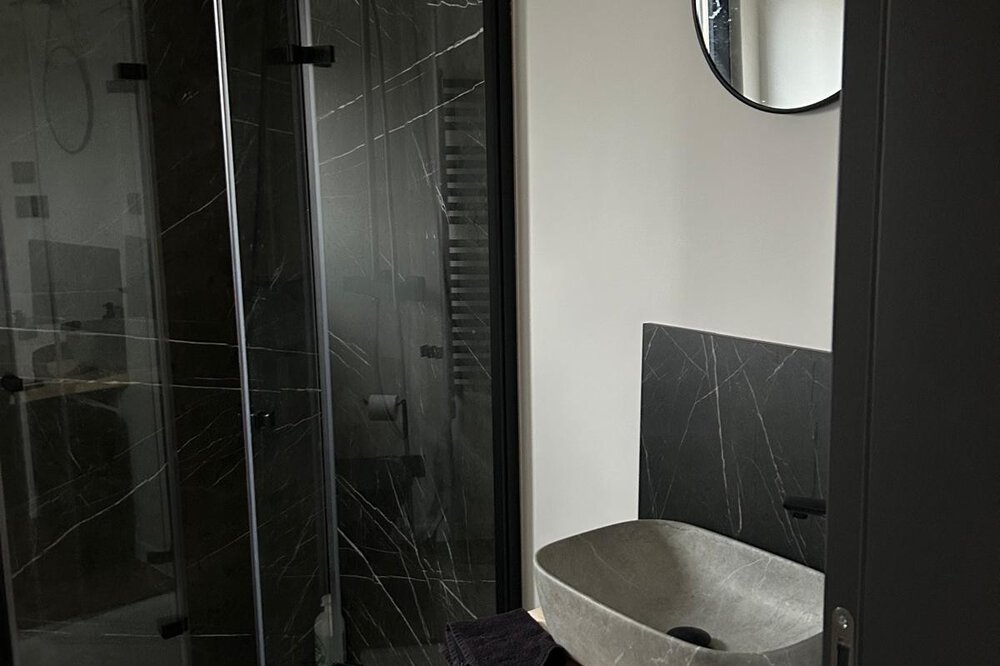 Modernes Badezimmer mit Glasdusche, rundem Spiegel, grauem Steinwaschbecken und schwarzen Akzenten.