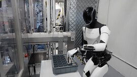 In a laboratory environment, a white humanoid robot sorts metal cylinders into a box.