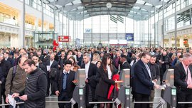 Zahlreiche Besucher stehen in der Eingangshalle der productronica Schlange, um ihre Tickets an den Drehkreuzen abzuscannen und einzutreten.