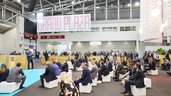 A large group of people sit on white seating cubes in an exhibition hall and listen to a speaker.
