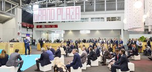 Eine große Gruppe von Menschen sitzt in einer Messehalle auf weißen Sitzwürfeln und lauscht einem Redner.