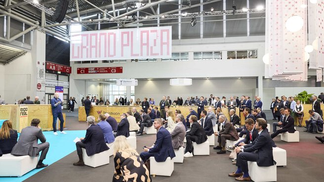A large group of people sit on white seating cubes in an exhibition hall and listen to a speaker.