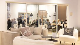 Gemütlicher Loungebereich mit Sofas und Kissen im Vordergrund, dahinter Tagungsraum mit mehreren Personen bei Gesprächen und Meetings an Tischen.