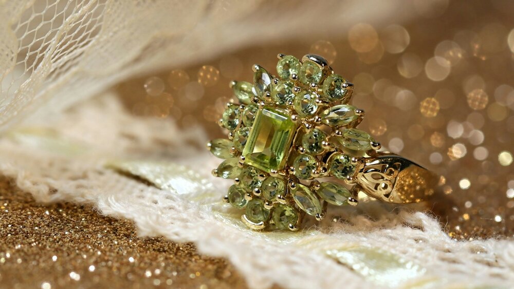 An ornate gold ring, set with numerous green gemstones arranged in the shape of a flower, rests on shimmering fabric.