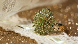 An ornate gold ring, set with numerous green gemstones arranged in the shape of a flower, rests on shimmering fabric.