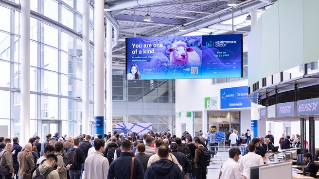 The entrance hall of the Kölnmesse, with visitors waiting in front of the turnstile. A digital screen hangs from the ceiling, displaying a cartoon sheep and the message “you are one of a kind”