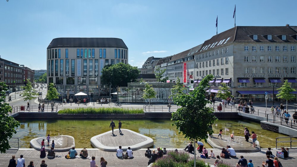  A lively city square with water features, green recreational areas, and surrounding modern and historic buildings, where people gather on sunny days.