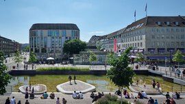  A lively city square with water features, green recreational areas, and surrounding modern and historic buildings, where people gather on sunny days.