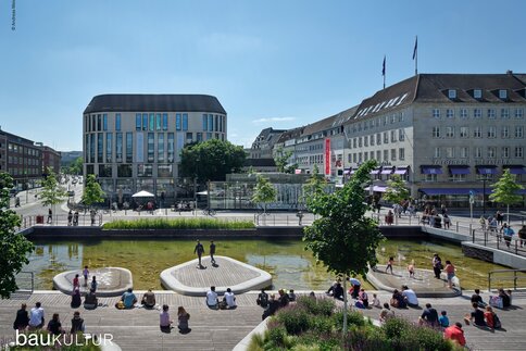 Belebter Stadtplatz mit Wasserflächen, begrünten Aufenthaltsbereichen und umliegenden modernen sowie historischen Gebäuden, auf dem sich Menschen bei sonnigem Wetter aufhalten.