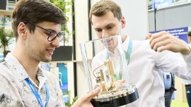 Zwei junge Männer betrachten auf der World of Quantum in München ein technisches Exponat in einer Glasvitrine mit großem Interesse.