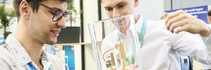 Zwei junge Männer betrachten auf der World of Quantum in München ein technisches Exponat in einer Glasvitrine mit großem Interesse.