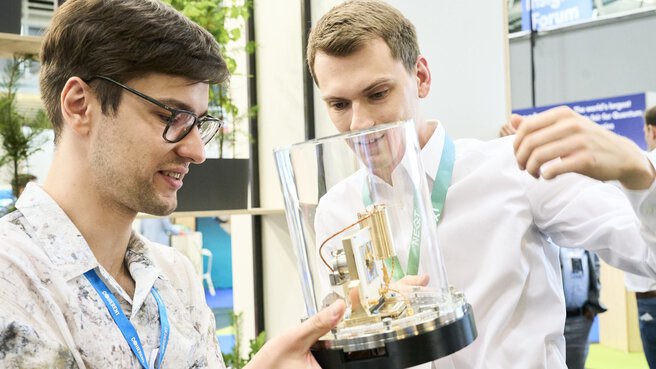 Zwei junge Männer betrachten auf der World of Quantum in München ein technisches Exponat in einer Glasvitrine mit großem Interesse.