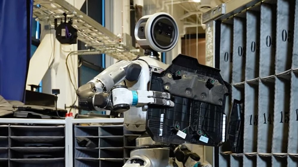 Ein humanoider Roboter steht vor einem Regal mit nummerierten Fächern und hält ein schwarzes Bauteil in der Hand.