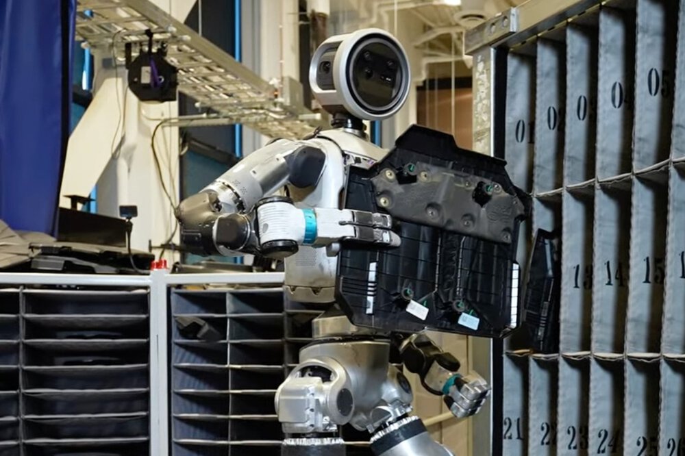 A humanoid robot stands in front of a shelf with numbered compartments and holds a black component in its hand.