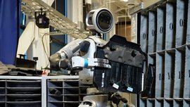 A humanoid robot stands in front of a shelf with numbered compartments and holds a black component in its hand.