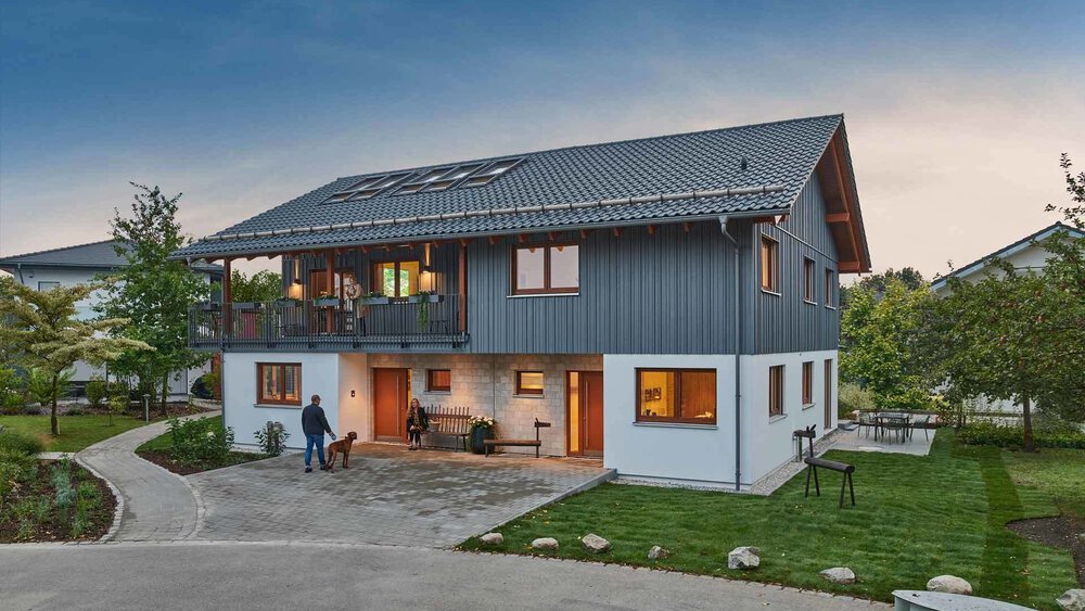 Modernes zweistöckiges Haus mit schrägem Dach, großen Fenstern und umgebendem Grün in der Abenddämmerung.