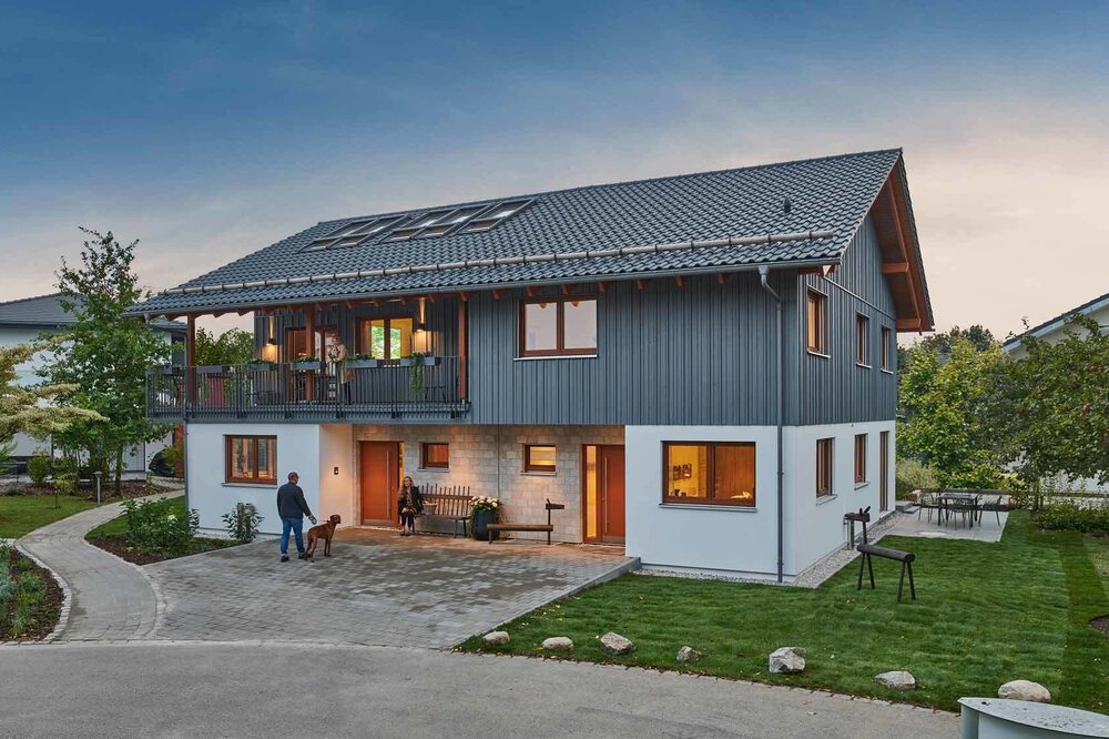 Modernes zweistöckiges Haus mit schrägem Dach, großen Fenstern und umgebendem Grün in der Abenddämmerung.