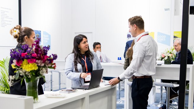 People are chatting at a trade fair booth; a laptop and flowers are on the white counter.