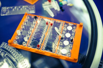 Close-up of a laboratory microplate with wells on an orange holder with blurred laboratory equipment in the background.