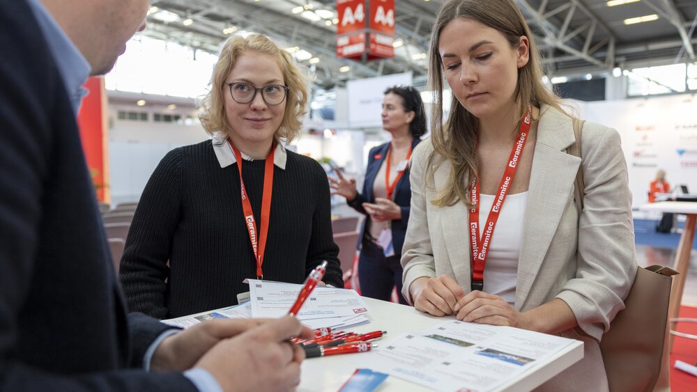 Two women are standing at an information stand at ceramitec and talking to an exhibitor at a standing table.
