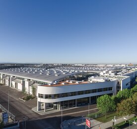 Ein großes, modernes Industriegebäude mit einem geschwungenen Dach, umgeben von Bäumen, unter einem strahlend blauen Himmel.