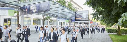 People in business attire walk on a paved path under a glass roof, surrounded by trees and buildings.