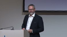 Ein dunkel gekleideter Mann mit Bart und Brille spricht am Rednerpult.