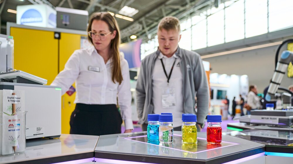 Two people are looking at colored liquid samples and lab equipment at a stand at a trade fair.