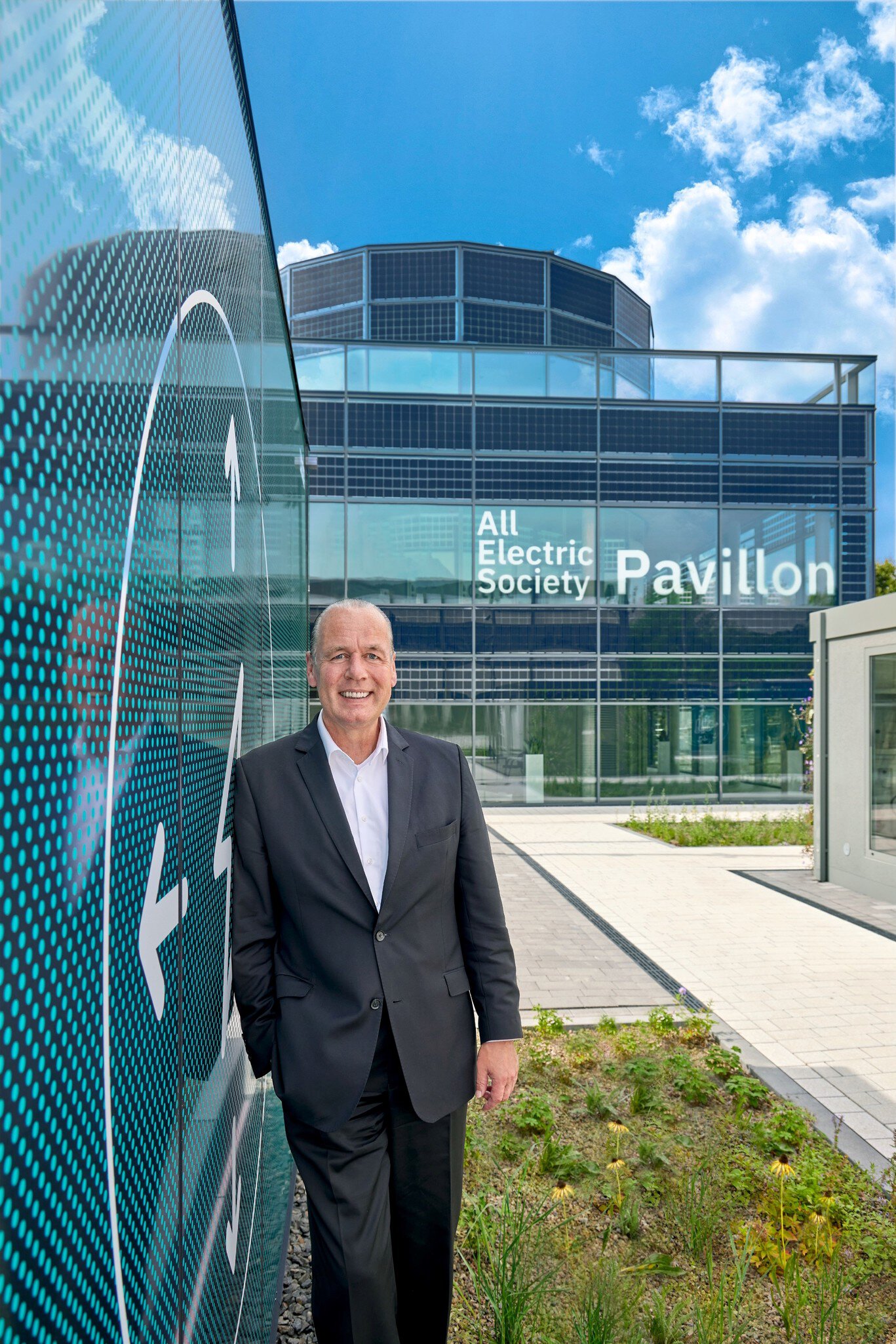 A man in a suit stands in front of a modern glass building with the inscription 