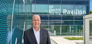 A man in a suit stands in front of a modern glass building with the inscription 