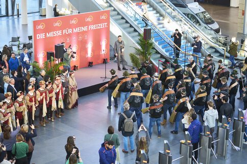 Blick aus der Vogelperspektive auf die Eröffnungsveranstaltung einer Messe in der Eingangshalle. Links ist eine Bühne, vor der Bühne führt eine Gruppe ein traditionelle Vorstellung mit großen Glocken auf. Rechts von der Bühne stehen Personen mit Trompeten.