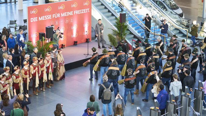 Bird's eye view of the opening event of a trade fair in the entrance hall. On the left is a stage, in front of the stage a group is performing a traditional show with large bells. To the right of the stage are people with trumpets.
