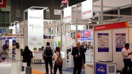 Several visitors walk around between numerous exhibition stands in an exhibition hall while the exhibitors wait.