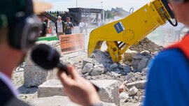 Close-up of a yellow hydraulic demolition shear crushing concrete elements, observed by trade fair visitors and a camera crew.