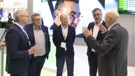 Fünf Männer in Anzügen diskutieren auf einer Konferenz, im Hintergrund ist ein großer Bildschirm zu sehen mit dem Gesichts eines Mannes mit Brille.