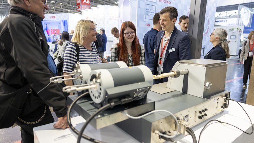 Mehrere Menschen unterhalten sich mit einem Aussteller vor einem ausgestellten elektrischen Gerät an einem Messestand.