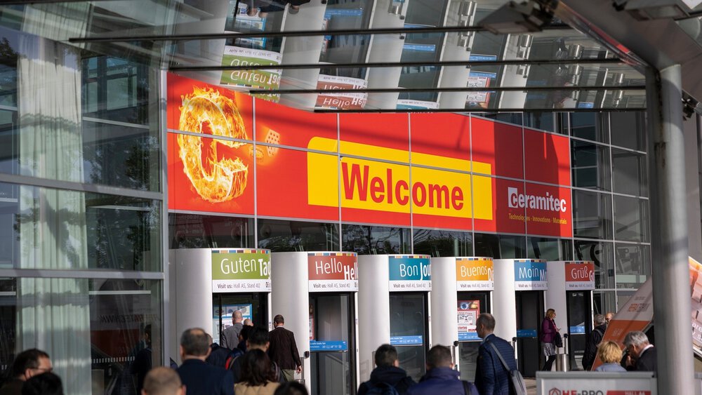 Several revolving doors leading to the entrance hall of the ceramitec trade fair, with a large red welcome sign above them and numerous people entering the building.